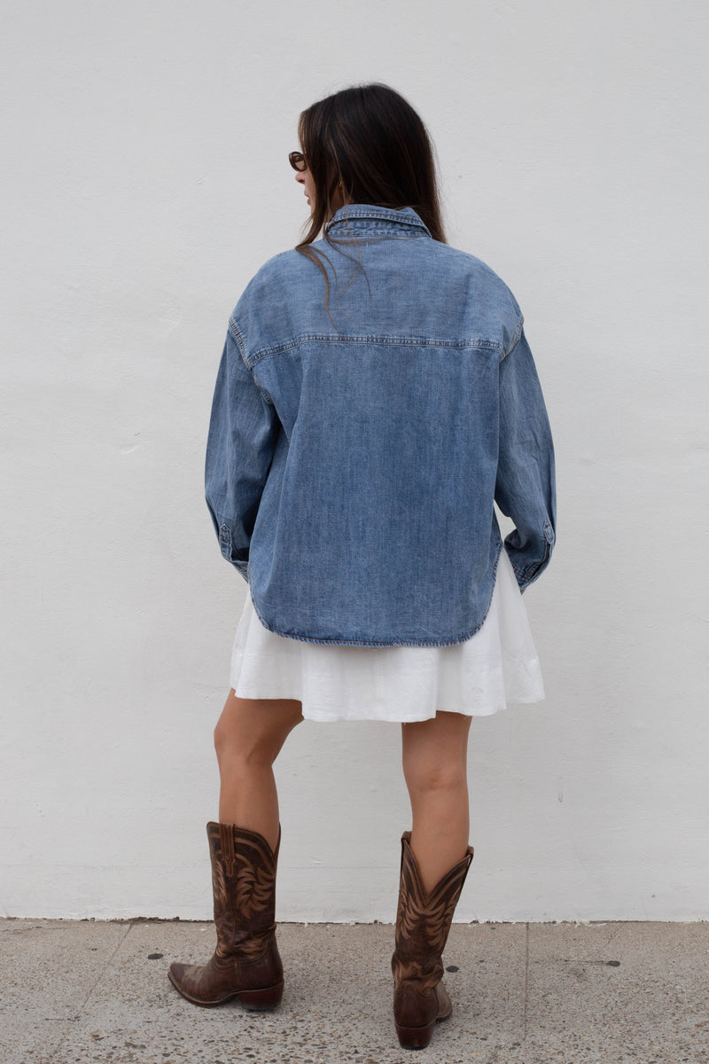 A woman shown from behind wearing an oversized blue denim shirt over a white dress, paired with brown cowboy boots, standing against a plain white wall.
