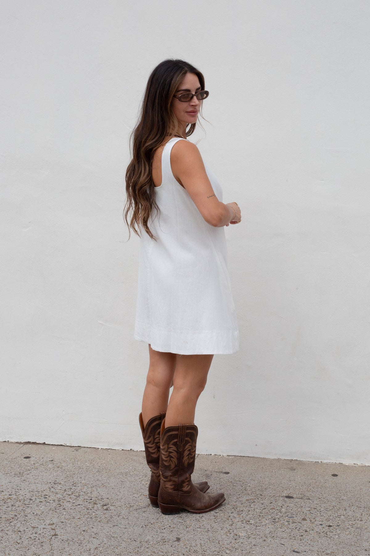 A woman wearing a sleeveless white mini dress and brown cowboy boots, standing against a plain white wall.