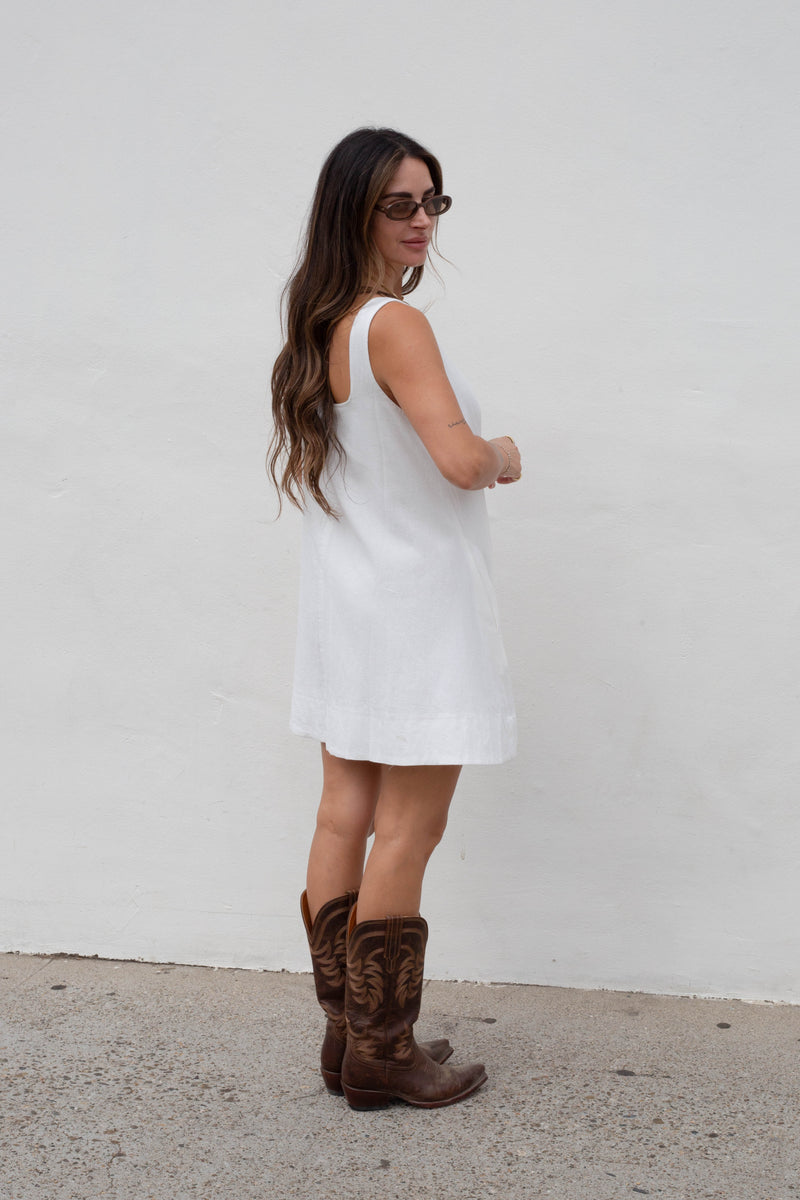 A woman wearing a sleeveless white mini dress and brown cowboy boots, standing against a plain white wall.