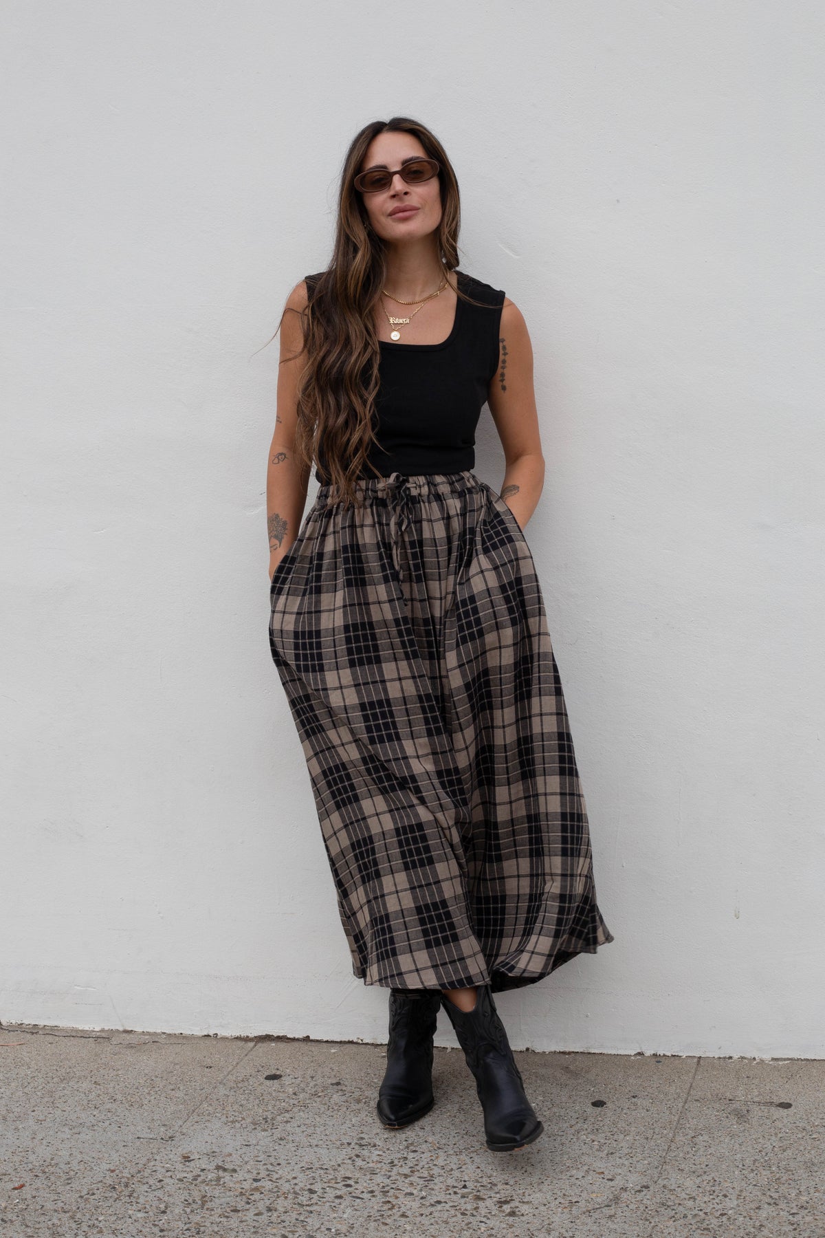 A woman stands against a white wall wearing a black sleeveless top, wide-leg plaid maxi skirt in brown and beige, and black ankle boots.