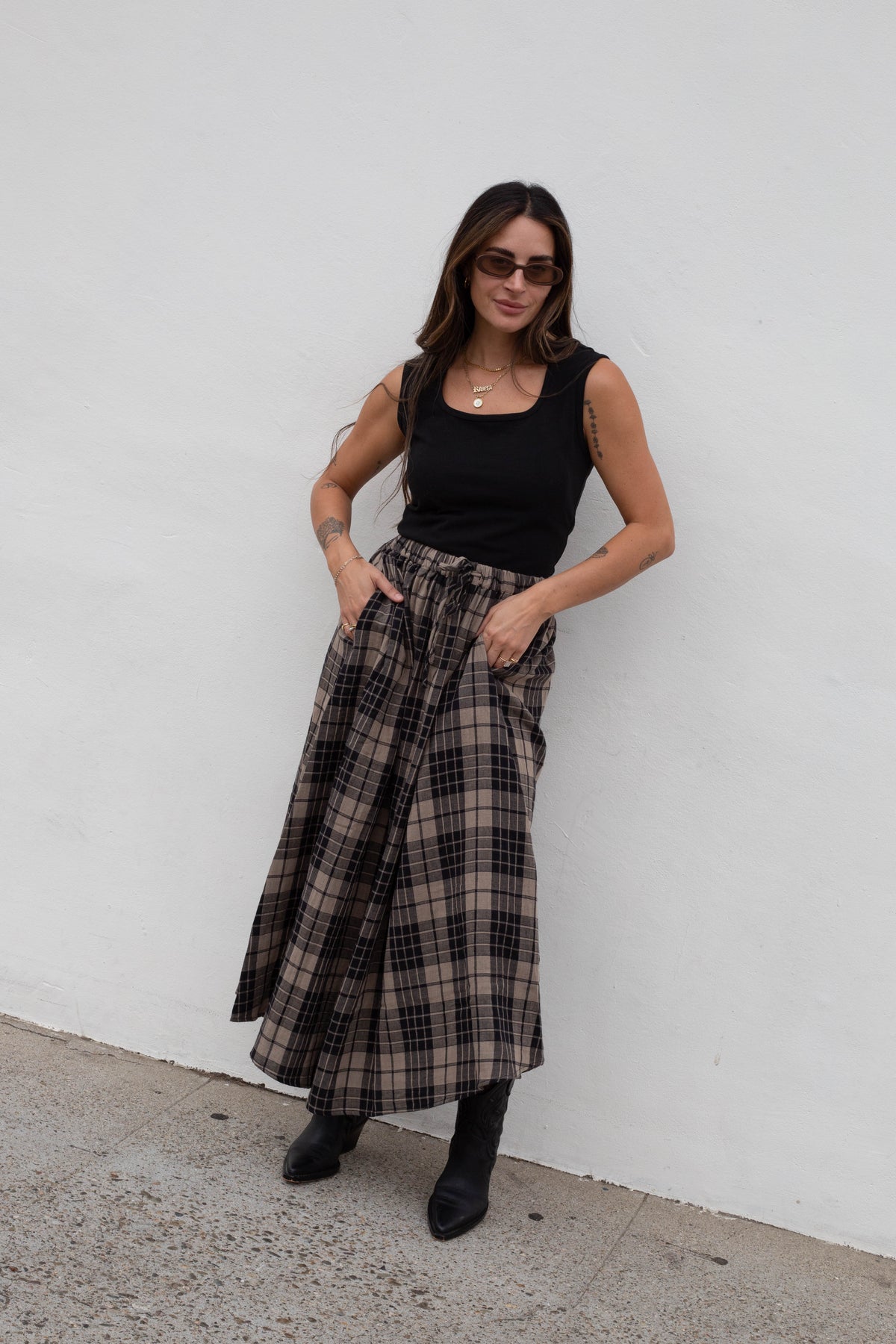 A woman wearing a black sleeveless top and a long brown and black plaid skirt, standing against a white wall.