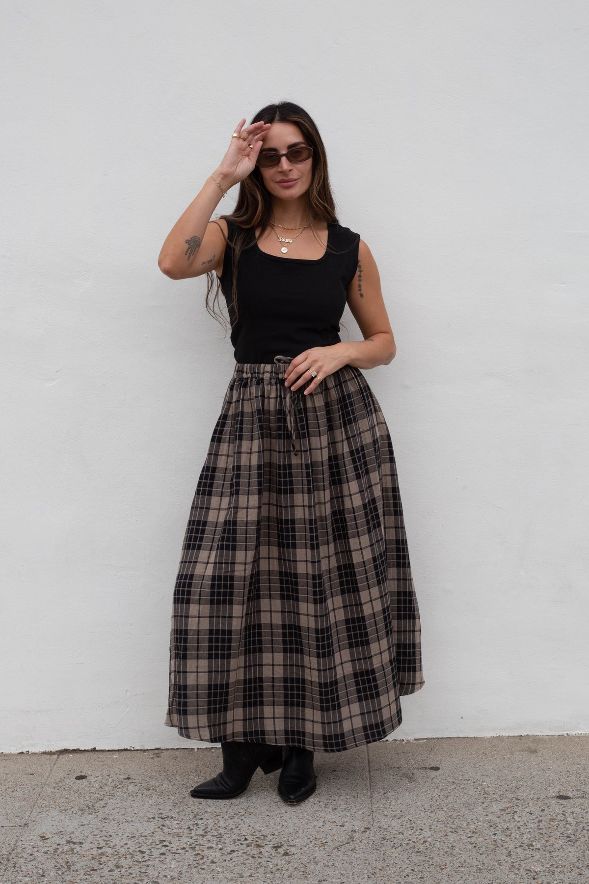 A woman stands against a white wall wearing a black sleeveless top and a long brown and black plaid skirt, paired with black boots.