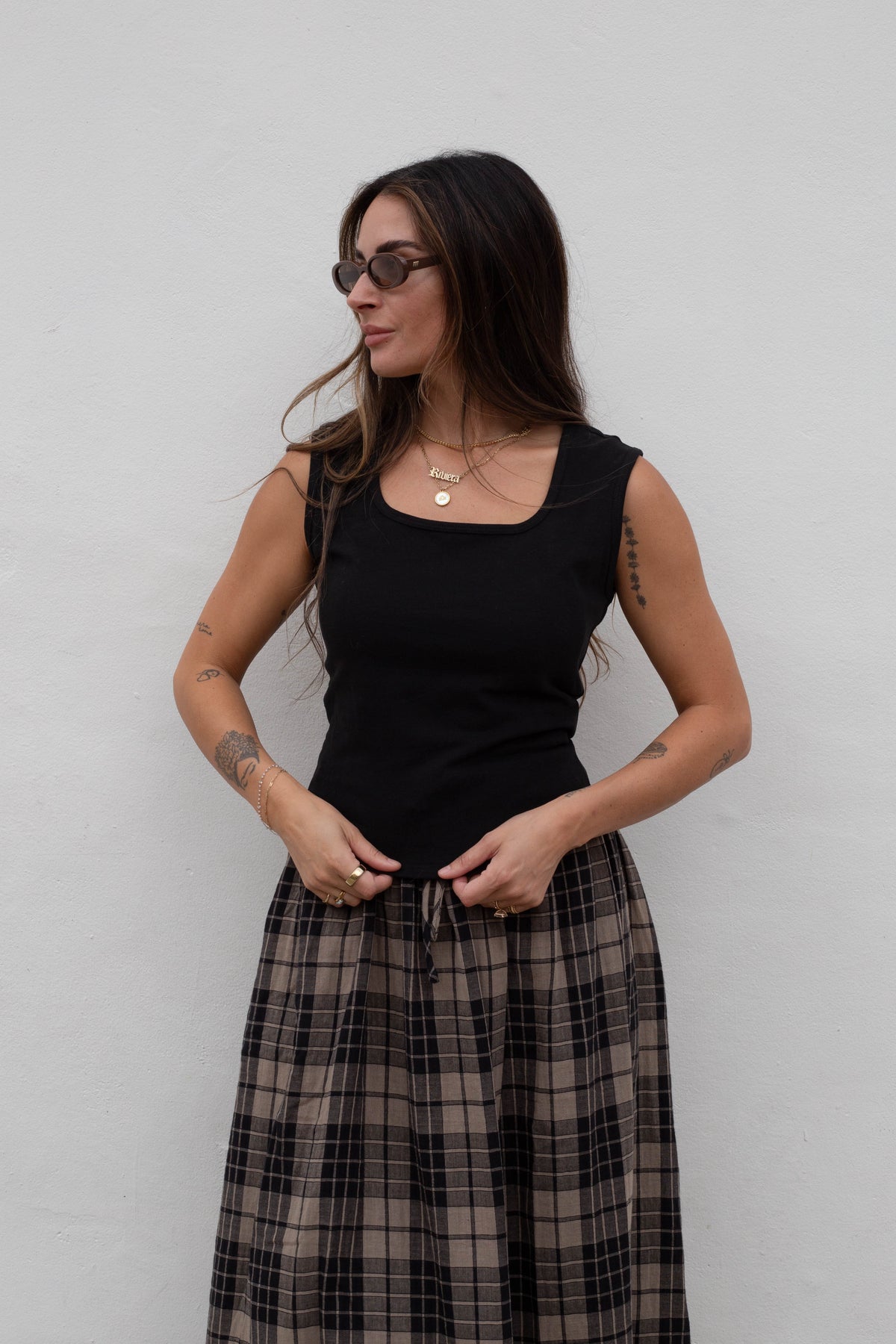 A woman modeling a black sleeveless tank top and a brown and black plaid maxi skirt, standing against a plain light gray wall.
