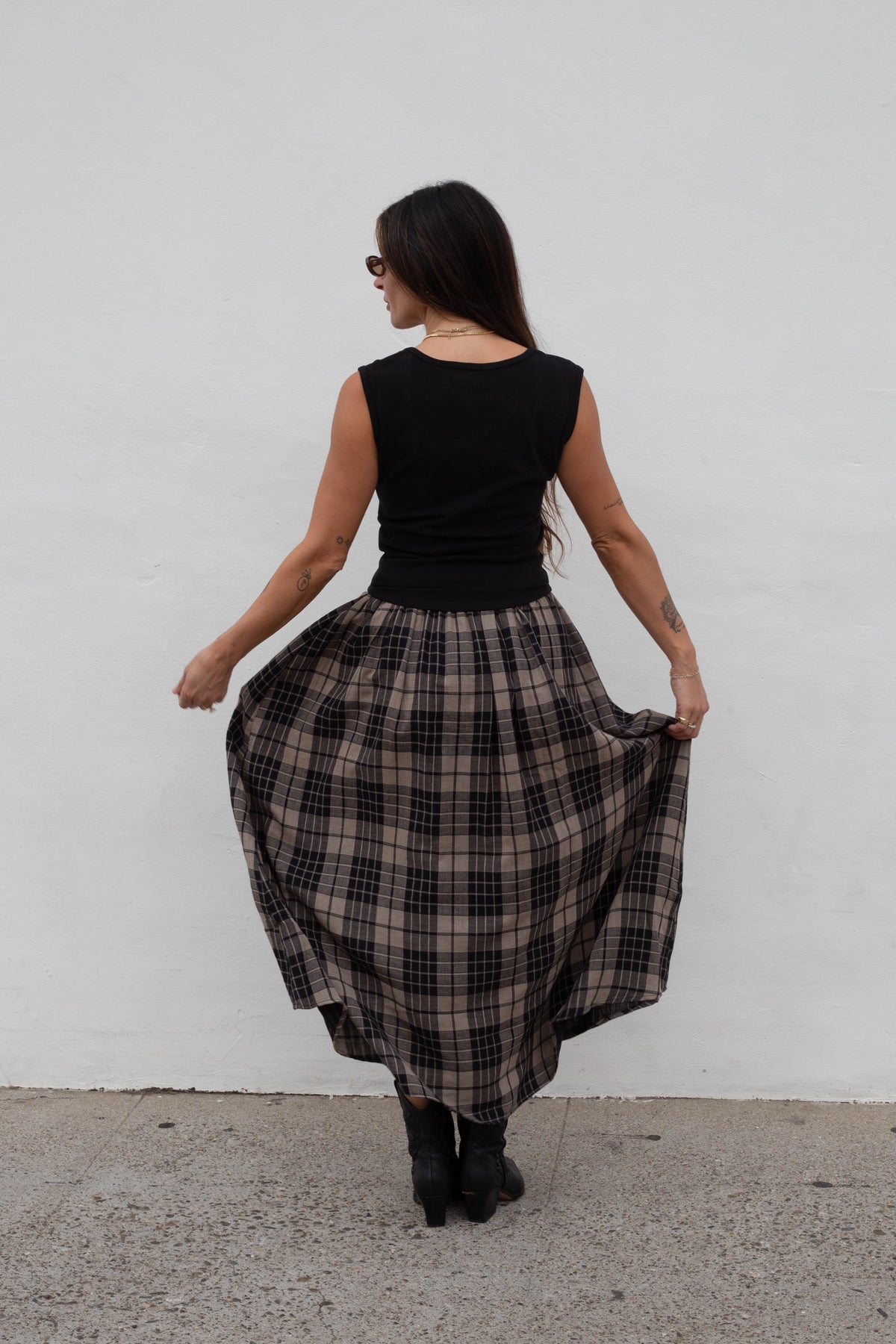 A woman is shown from behind wearing a sleeveless black top and a full black and beige plaid skirt, holding out the skirt to show its volume against a plain white wall.