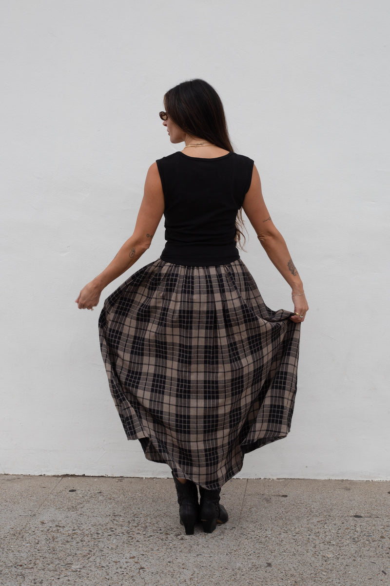 A woman is shown from behind wearing a sleeveless black top and a full black and beige plaid skirt, holding out the skirt to show its volume against a plain white wall.