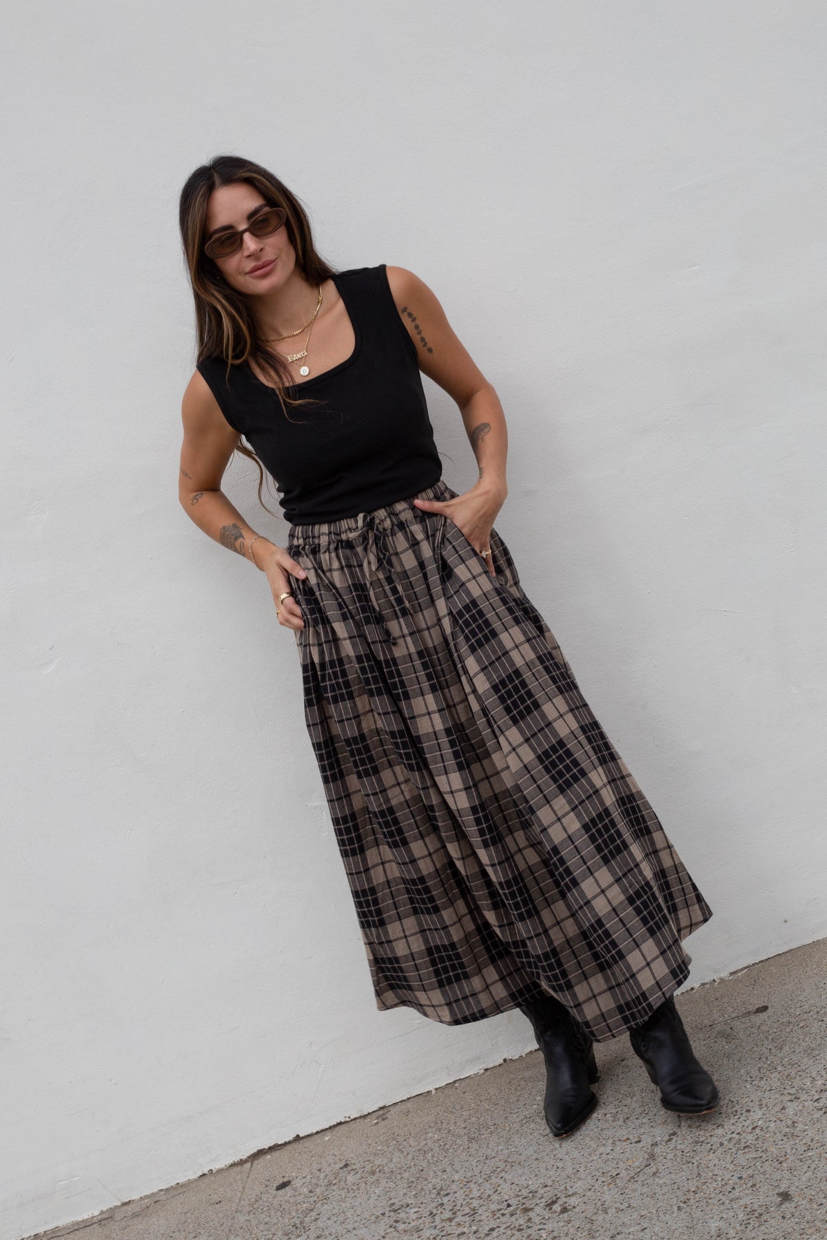 A woman wearing a black sleeveless top and a long brown and black plaid midi skirt, posing against a white wall.