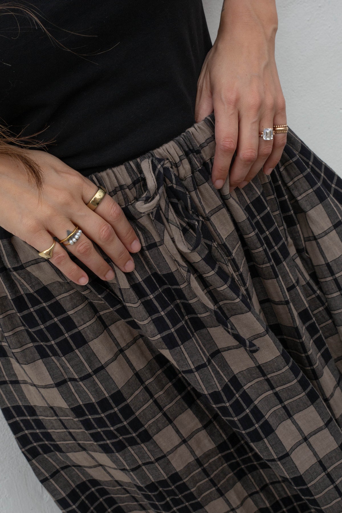Close-up of a person wearing wide-leg plaid pants in brown and black, with a black top and gold rings visible on their hands against a light gray wall.
