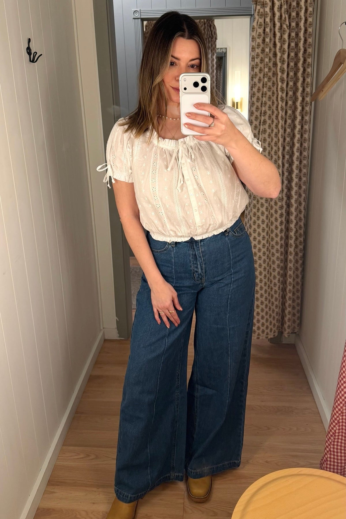Woman taking a mirror selfie wearing a white blouse and blue jeans in a room with a curtain and wooden floor.