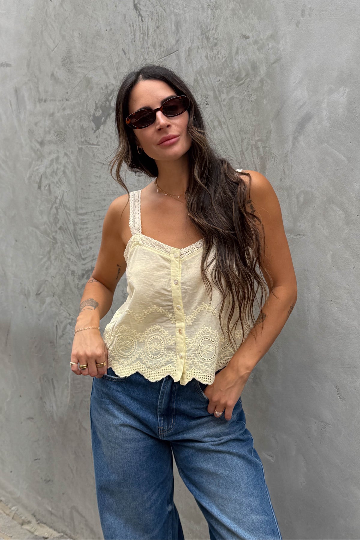 Woman wearing a yellow lace top and blue jeans against a gray wall.