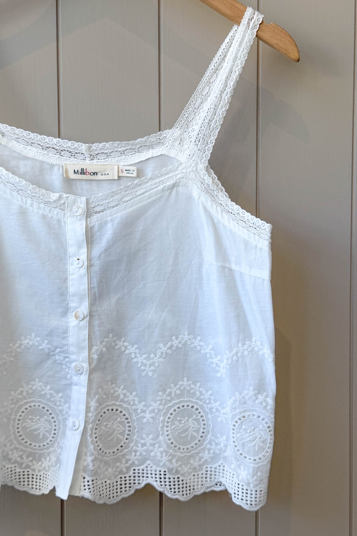 White sleeveless camisole top with lace trim and button front, hanging on a wooden hanger against a beige paneled wall.