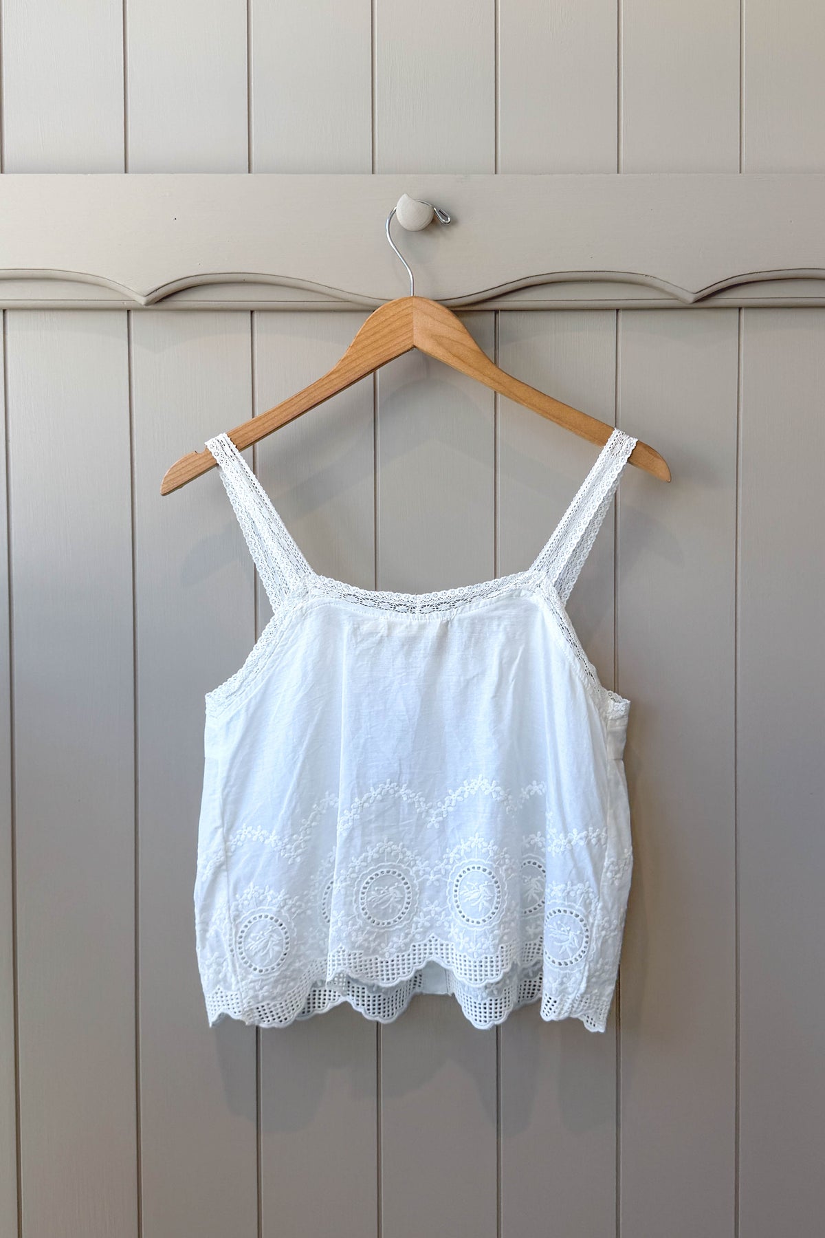 A white sleeveless lace-trim camisole top hanging on a wooden hanger against a light gray paneled wall.