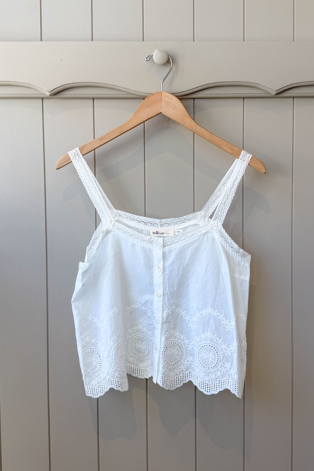 A white sleeveless lace-trim camisole top hanging on a wooden hanger against a light gray paneled wall.