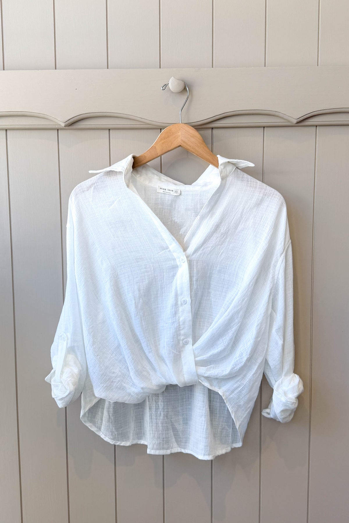 A white long-sleeve button-up shirt with a twisted front detail and rolled cuffs, hanging on a wooden hanger against a light gray paneled wall.