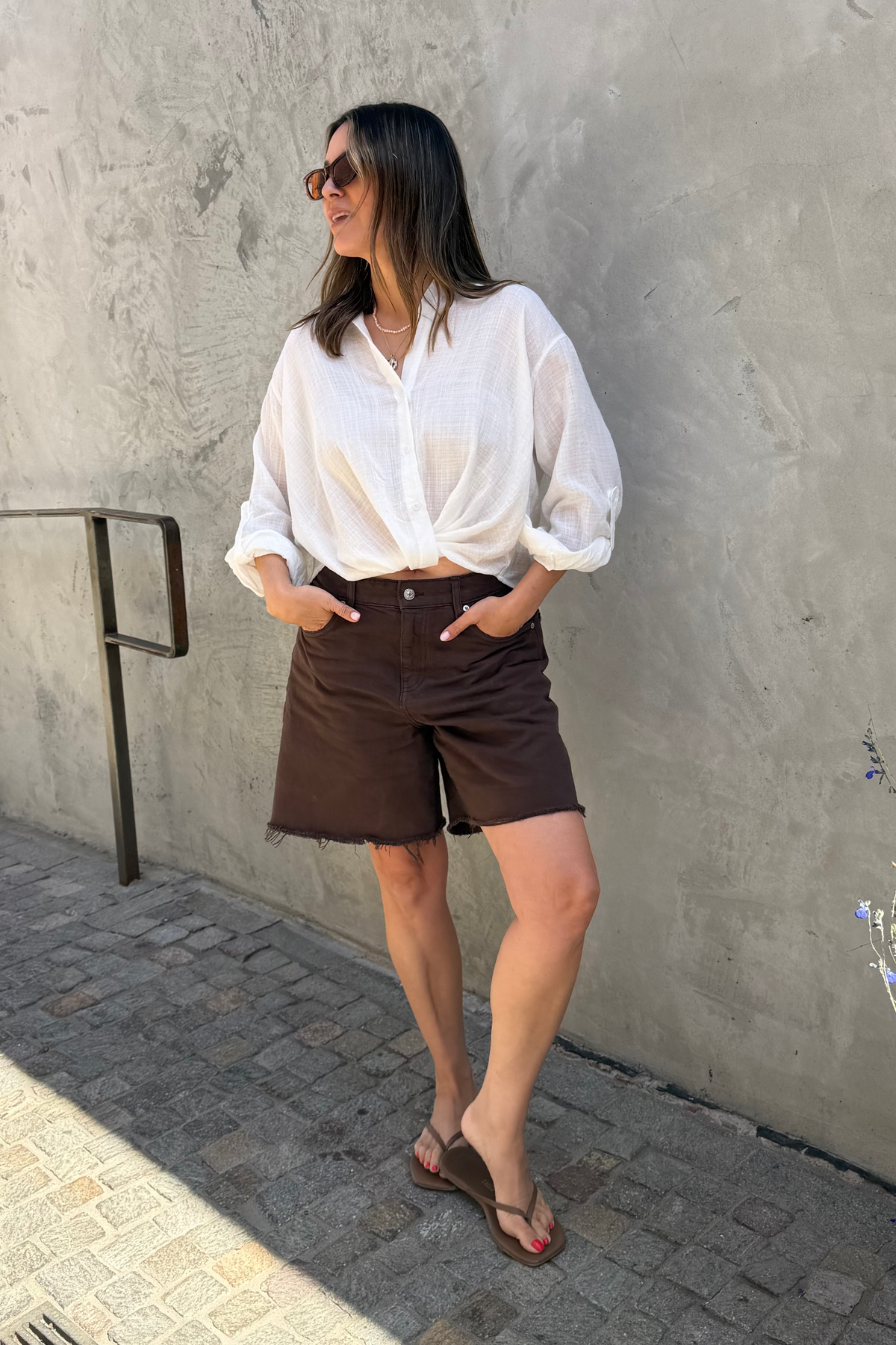 A woman stands outdoors against a textured concrete wall wearing a white long-sleeve button-up shirt, dark brown shorts, and brown sandals.