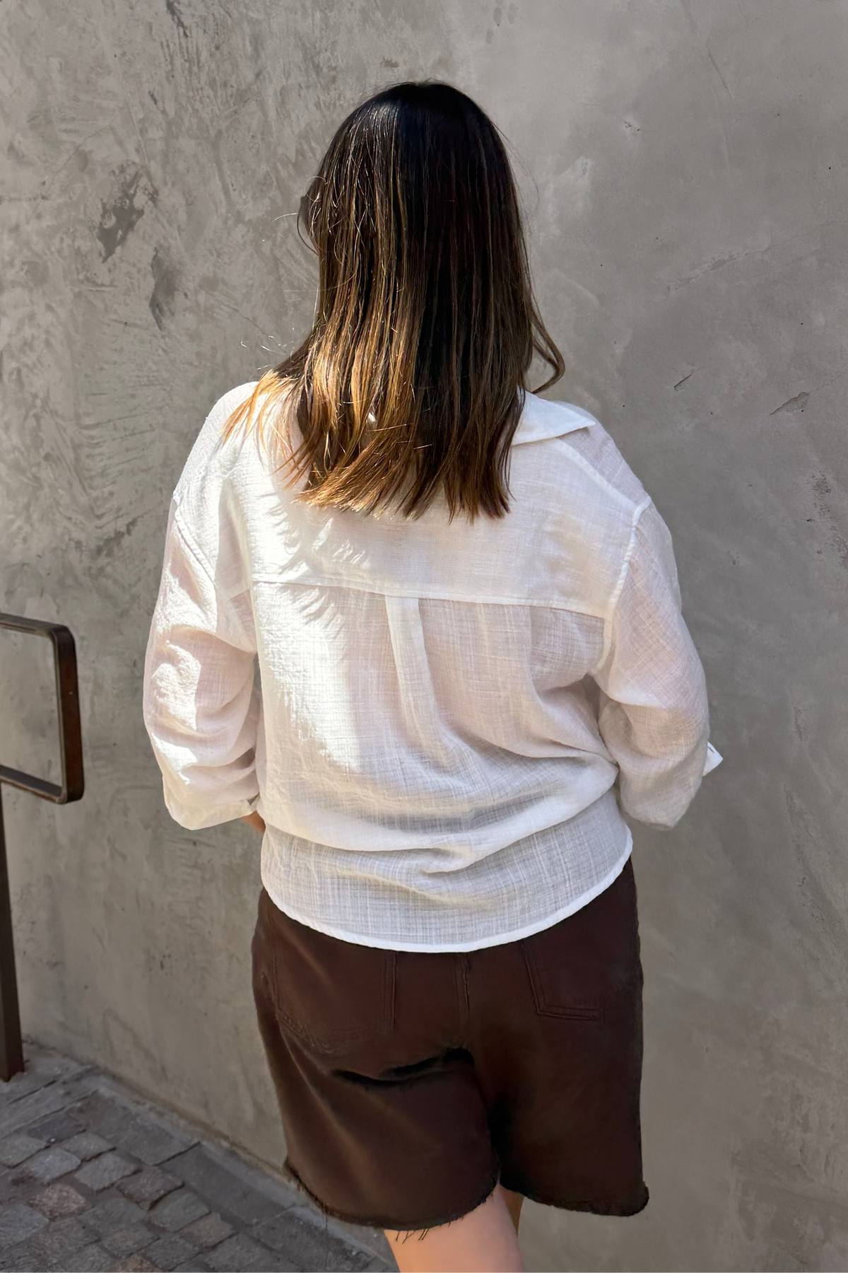 A woman viewed from behind wearing a white textured long-sleeve button-up shirt and dark brown shorts, standing against a textured gray concrete wall.