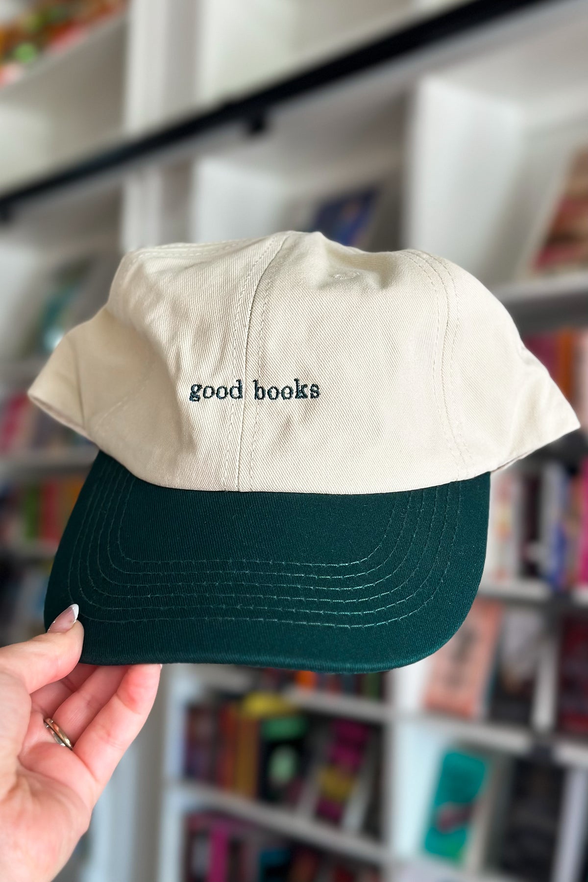Beige and green cap with 'good books' text held in front of a bookshelf.