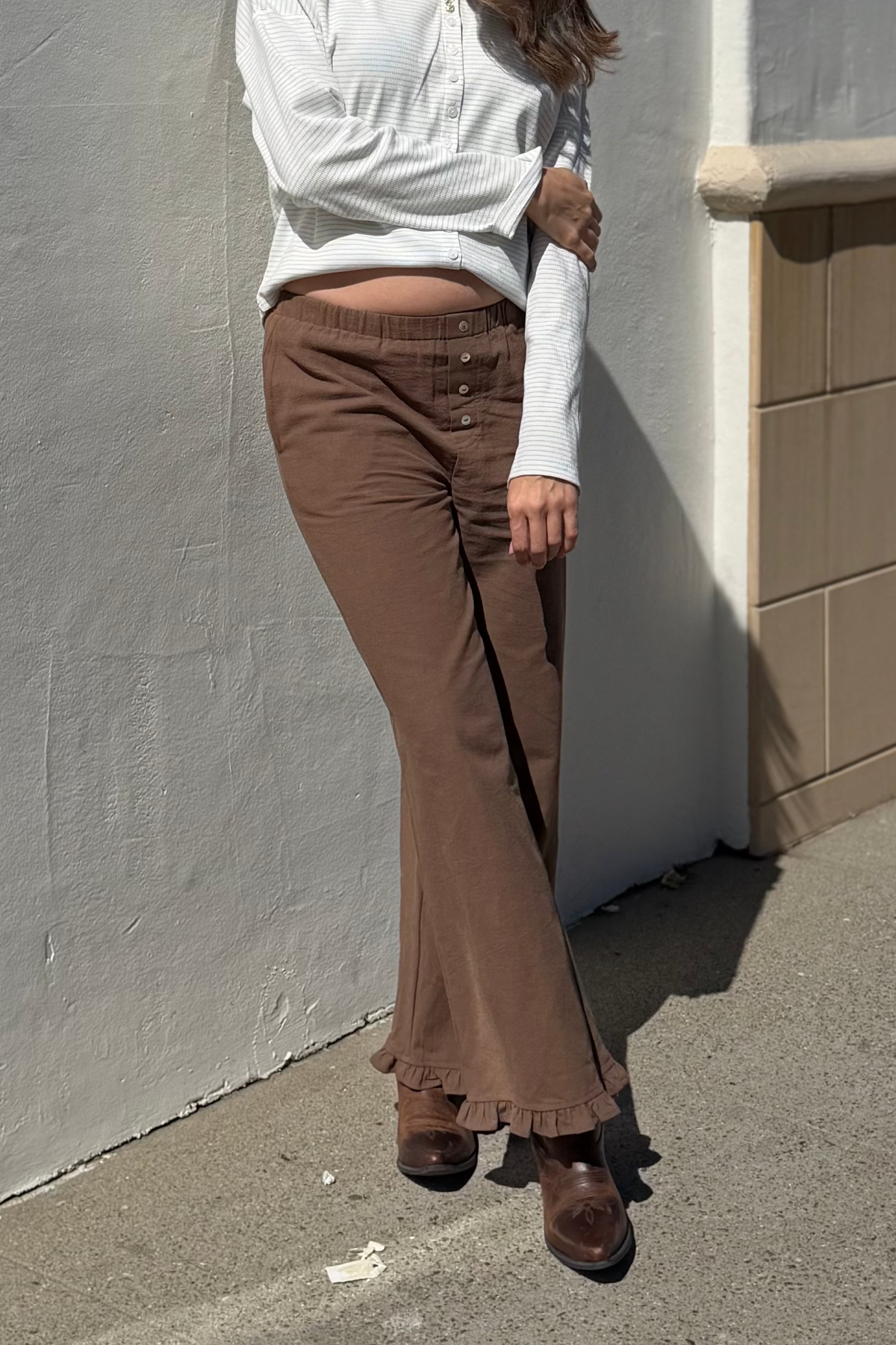 Woman in white top and brown pants standing against a gray wall.
