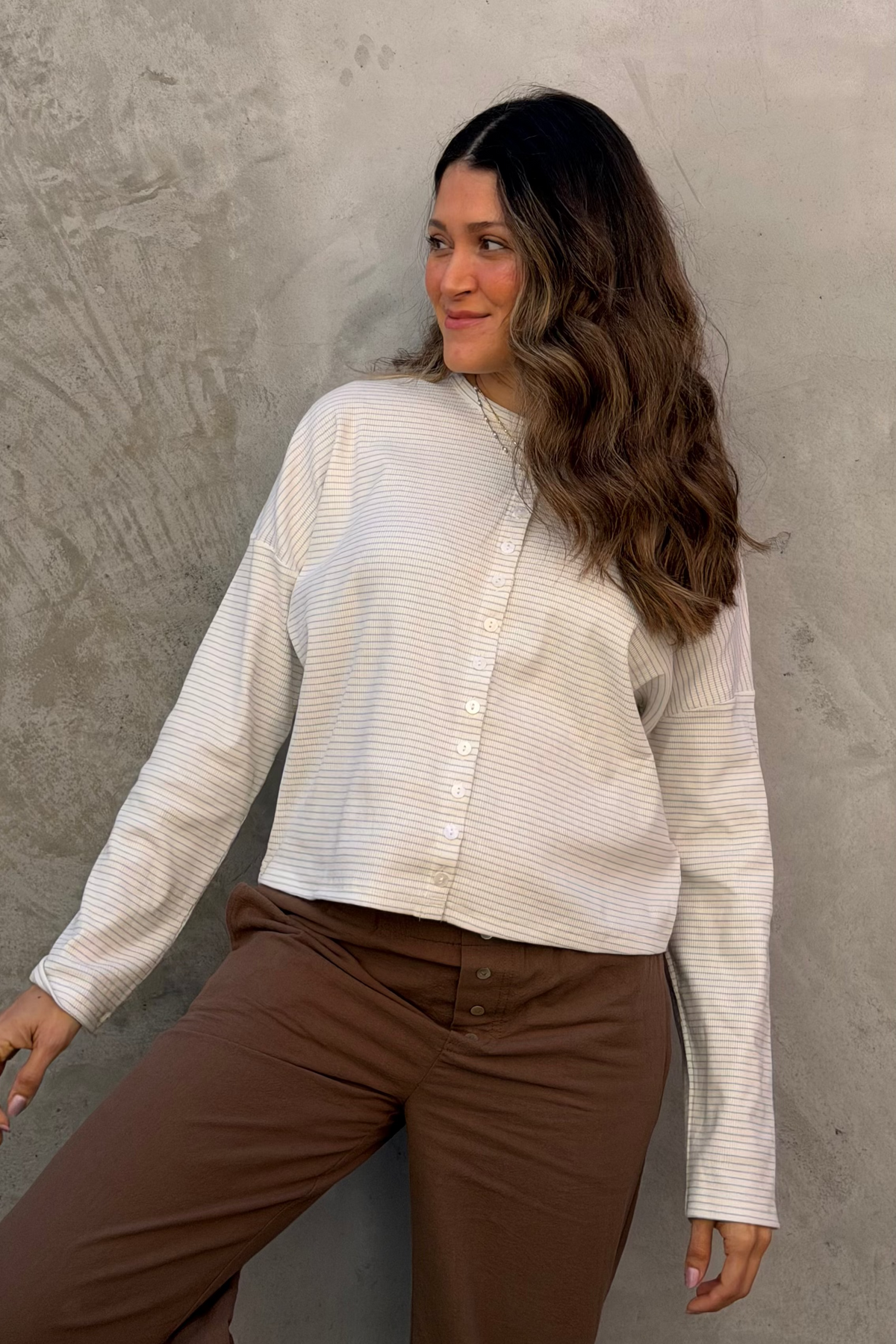 Woman wearing a white textured sweater and brown pants against a gray concrete wall.