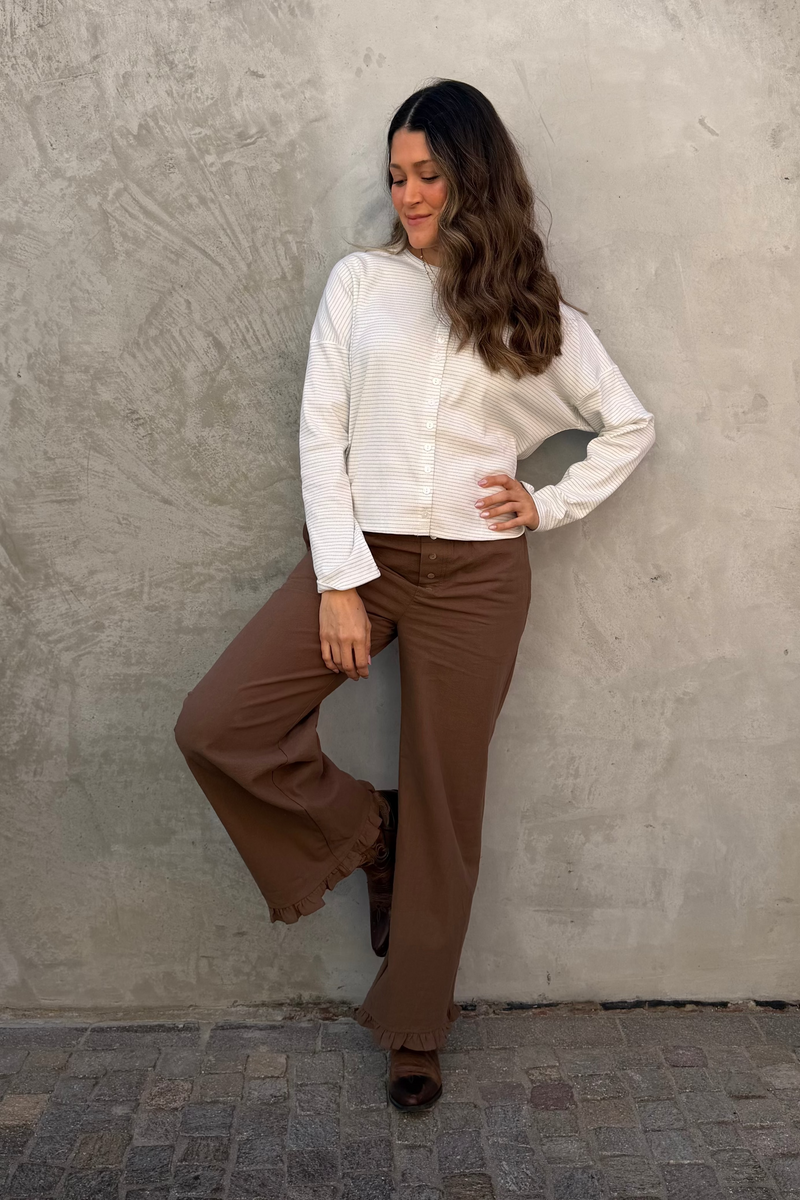 Woman in a white blouse and brown pants standing against a textured wall.