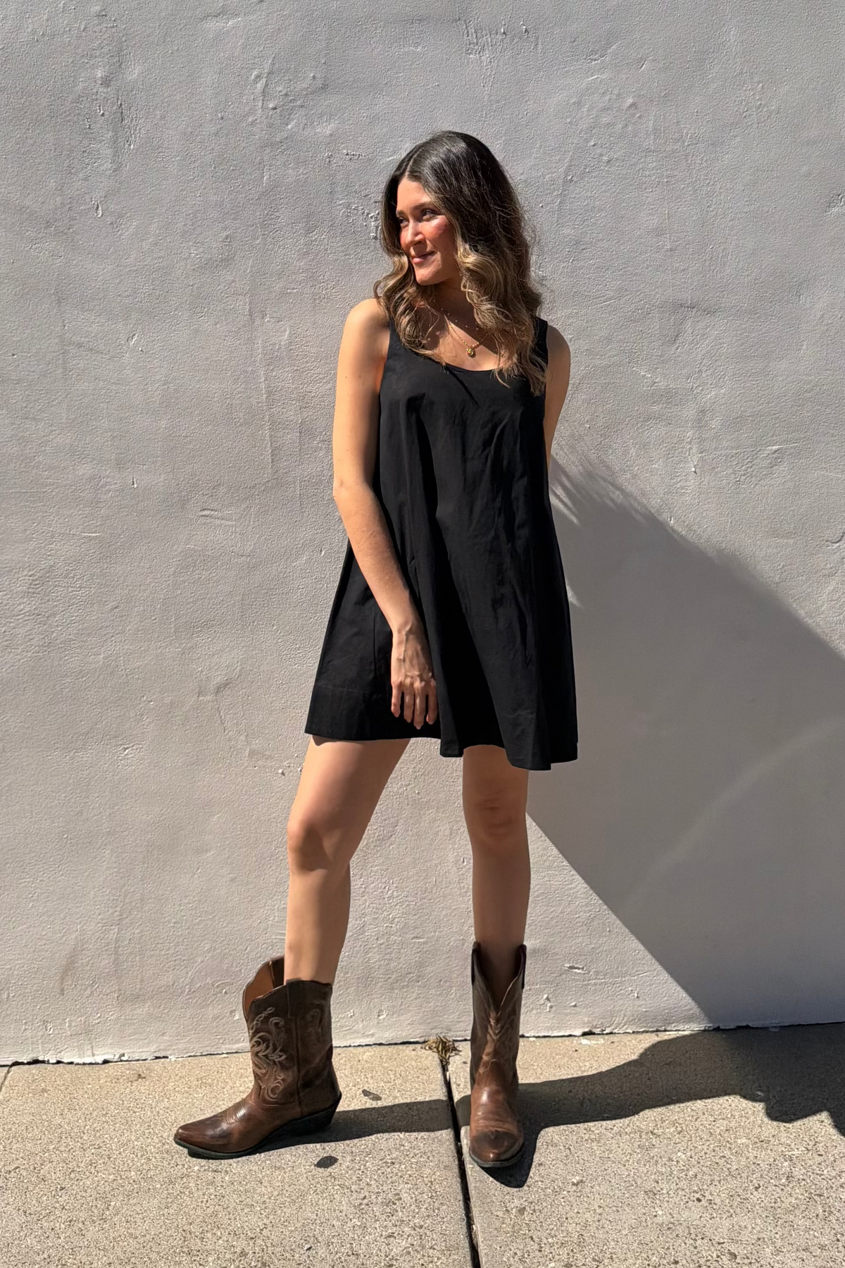Woman in a black dress and brown boots standing against a gray wall.