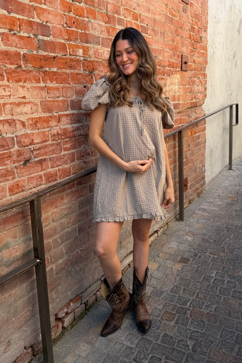Woman in a checkered dress and cowboy boots standing against a brick wall.