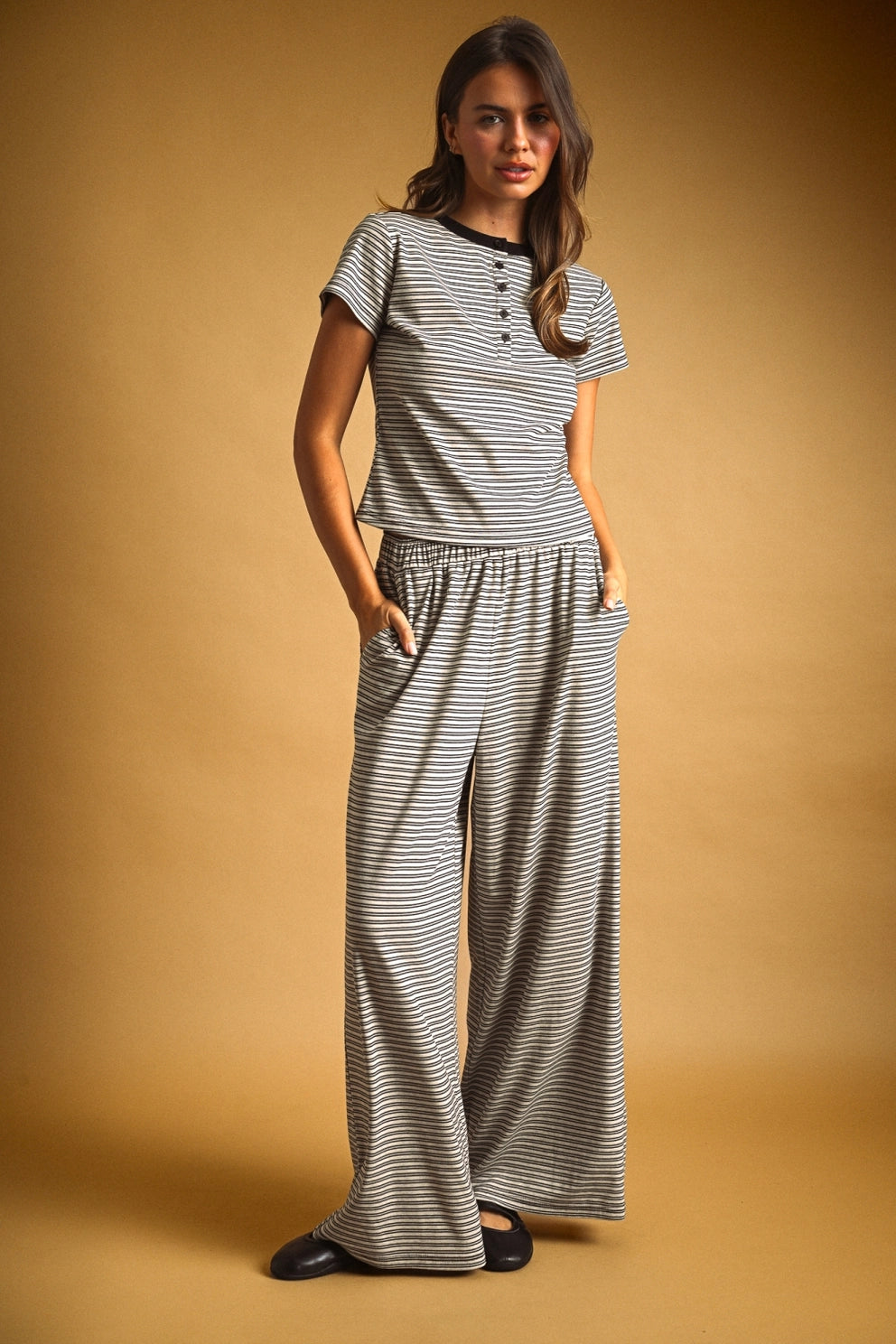 A woman modeling a matching two-piece loungewear set featuring a short-sleeve henley top and wide-leg pants, both in a gray and white striped pattern, set against a solid warm brown studio background.