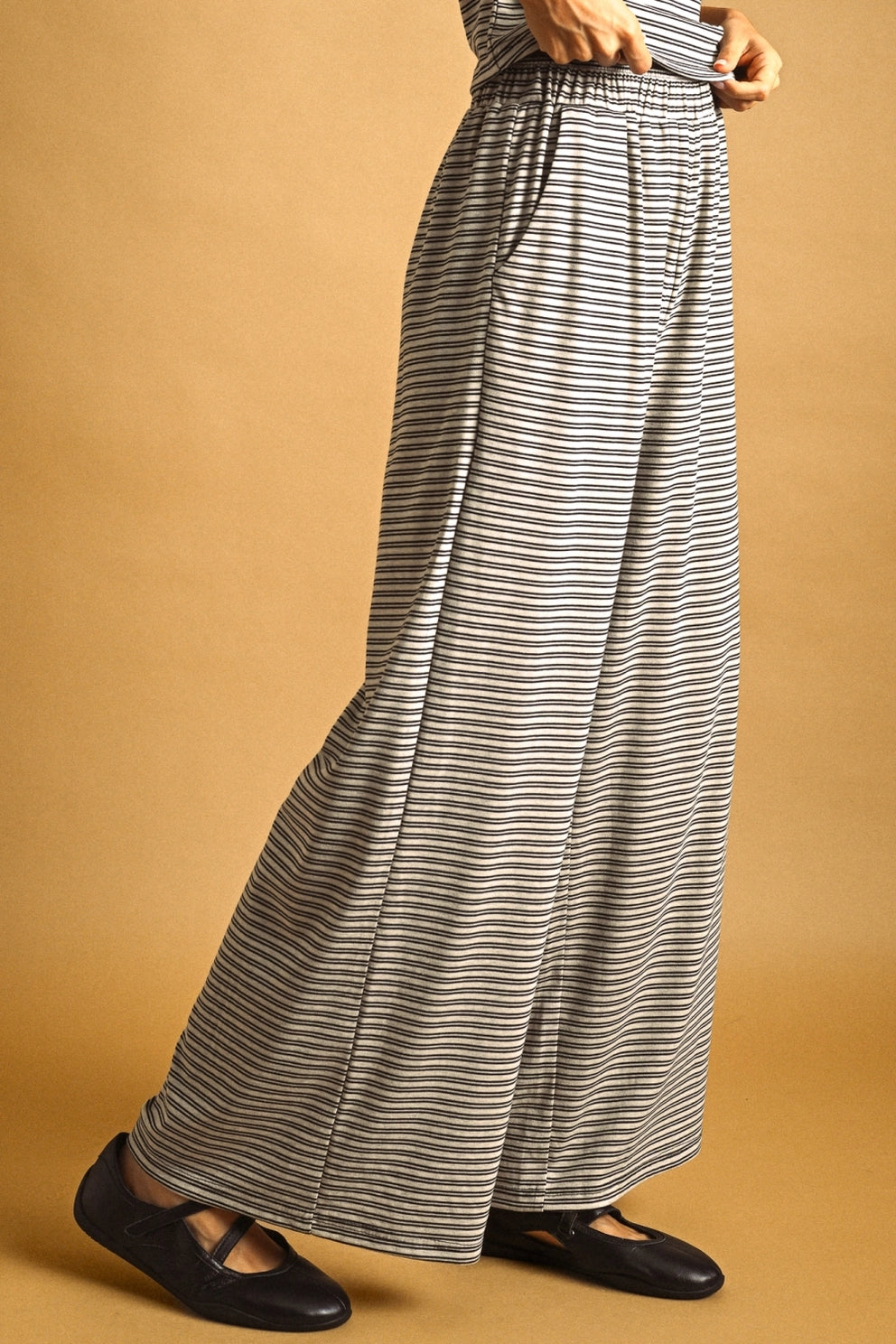 A person wearing wide-leg striped pants with a gray and white horizontal stripe pattern, paired with black flats, against a solid tan studio background.
