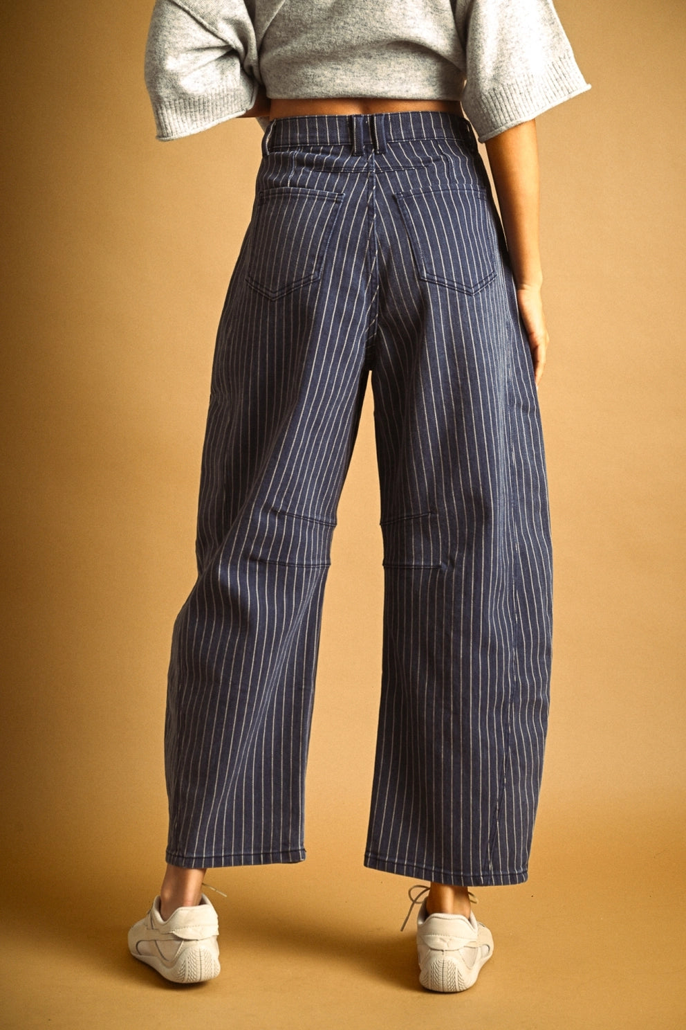 A person shown from the back wearing high-waisted, wide-leg cropped pants with thin vertical pinstripes, paired with a light gray knit top and white sneakers, against a warm tan studio backdrop.