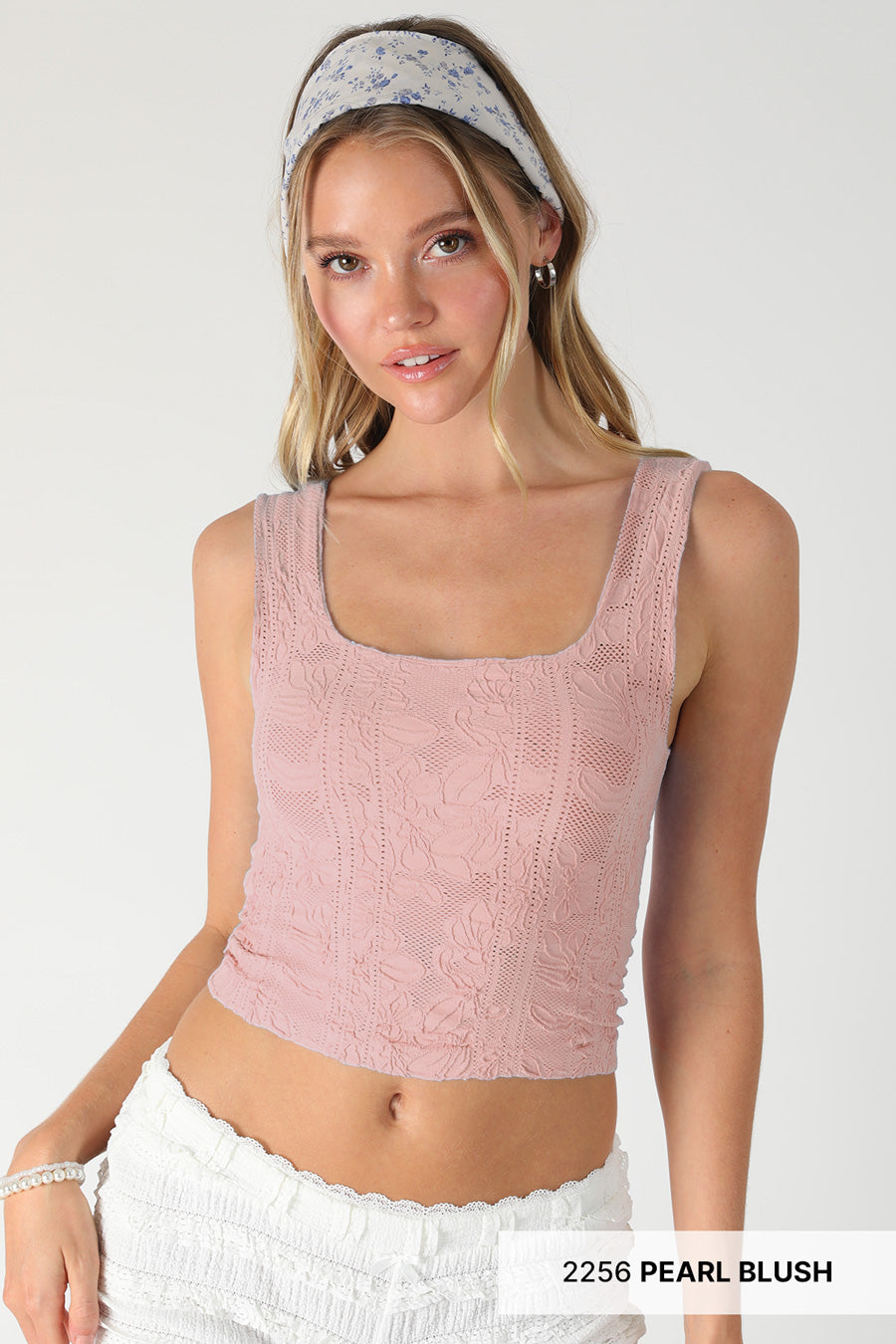 Woman wearing a pink knitted tank top with white pants on a plain background
