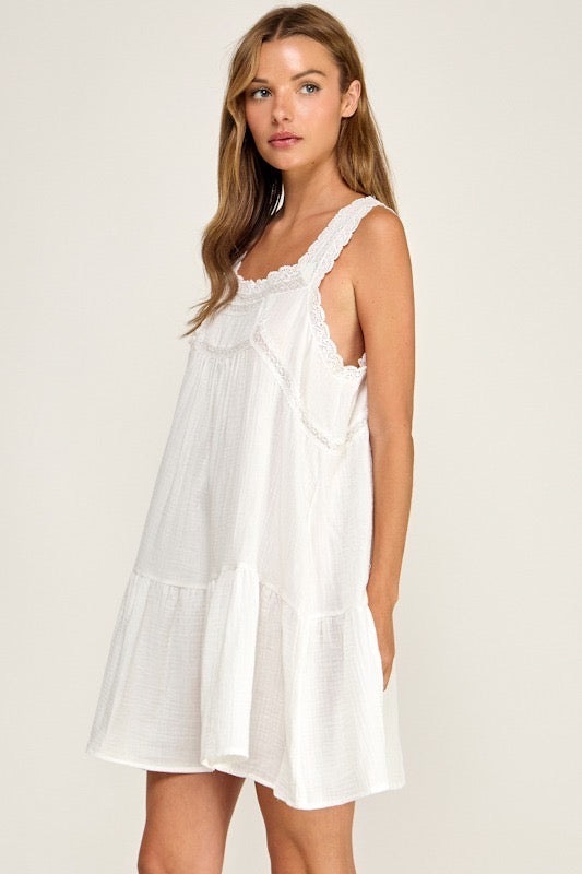 A woman modeling a white tiered cotton slip dress with ruffled trim and scalloped edges against a light beige studio background.