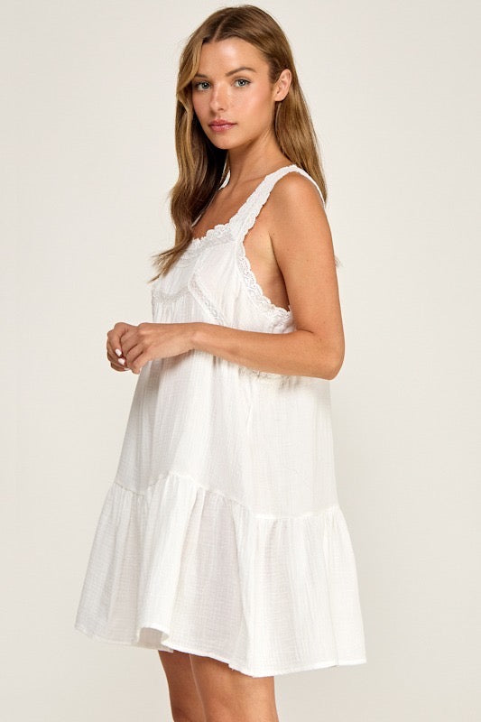 A woman wearing a white tiered sleeveless dress with lace trim, photographed against a plain light beige studio background.