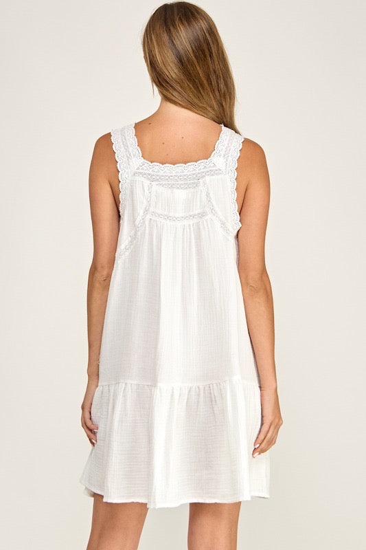 A woman shown from the back wearing a sleeveless white nightgown or short dress with lace trim at the neckline and a tiered hem, against a plain light beige studio background.