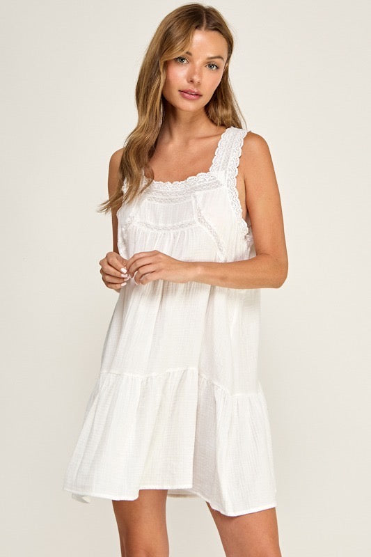 A woman modeling a white sleeveless tiered mini dress with lace trim on the neckline and shoulder straps, photographed against a plain light beige studio background.