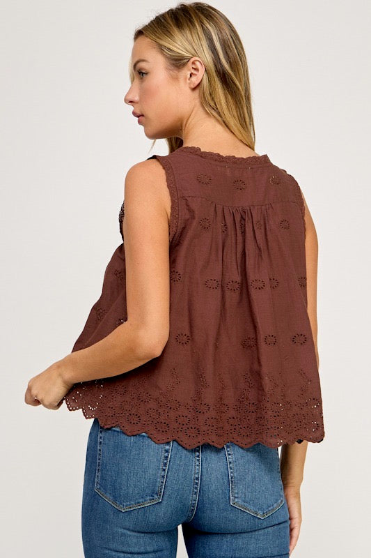 A woman shown from the back wearing a sleeveless brown eyelet lace top with a scalloped hem and blue jeans, against a light gray studio background.