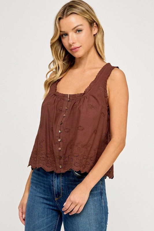 A woman wearing a brown sleeveless eyelet blouse with a button-down front and scalloped edges, paired with blue jeans, against a light gray studio background.