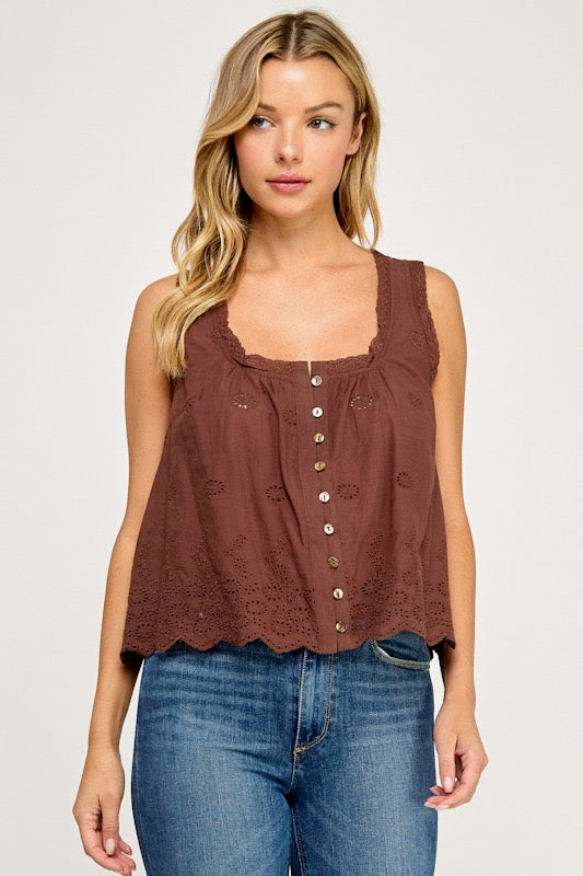 A woman wearing a brown sleeveless top with a scalloped eyelet hem and button-front detail, paired with blue jeans, against a light gray studio background.