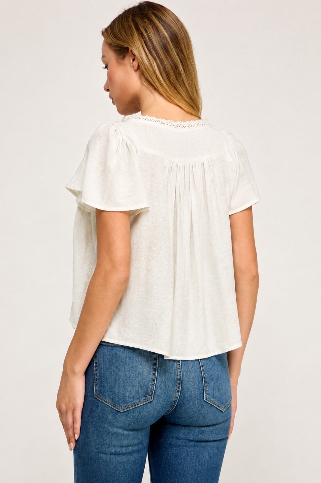 A woman shown from the back wearing a short-sleeve cream-colored blouse with delicate lace trim at the neckline and blue jeans, set against a plain light background.