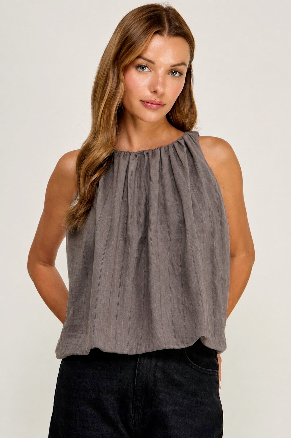A woman wearing a sleeveless, high-neck, gathered top in a textured gray fabric, paired with dark jeans, against a plain light gray studio background.