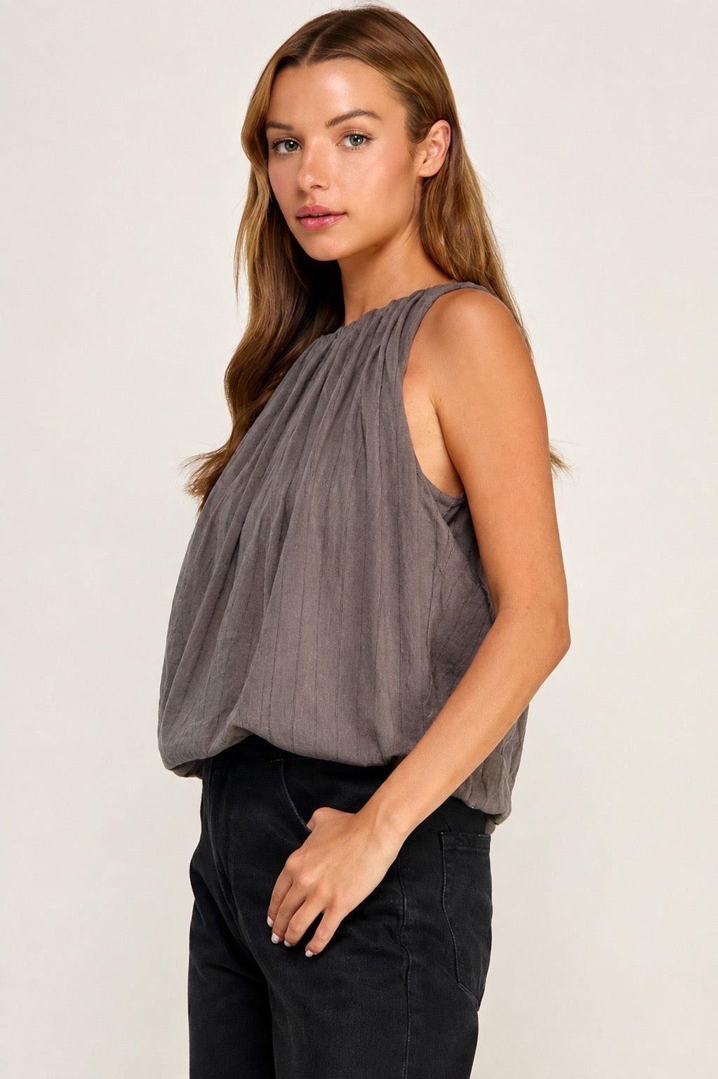A woman modeling a sleeveless, draped gray top paired with black pants, photographed against a plain light background.