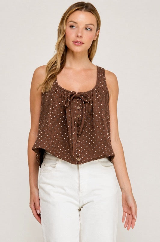 A woman modeling a brown sleeveless top with small white polka dots and a front tie detail, paired with white pants, against a plain light background.