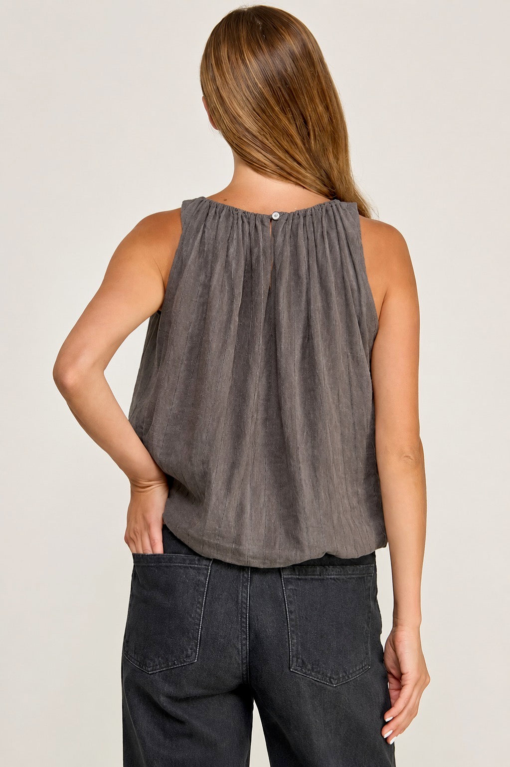 A woman shown from the back wearing a sleeveless dark gray top with a gathered neckline and dark jeans, against a plain light background.