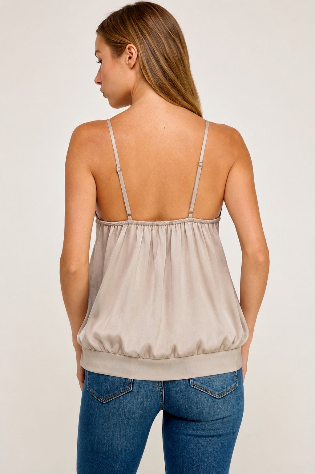 A woman shown from the back wearing a light beige spaghetti-strap camisole top with a gathered elastic waistband, paired with blue jeans, against a plain light gray studio background.