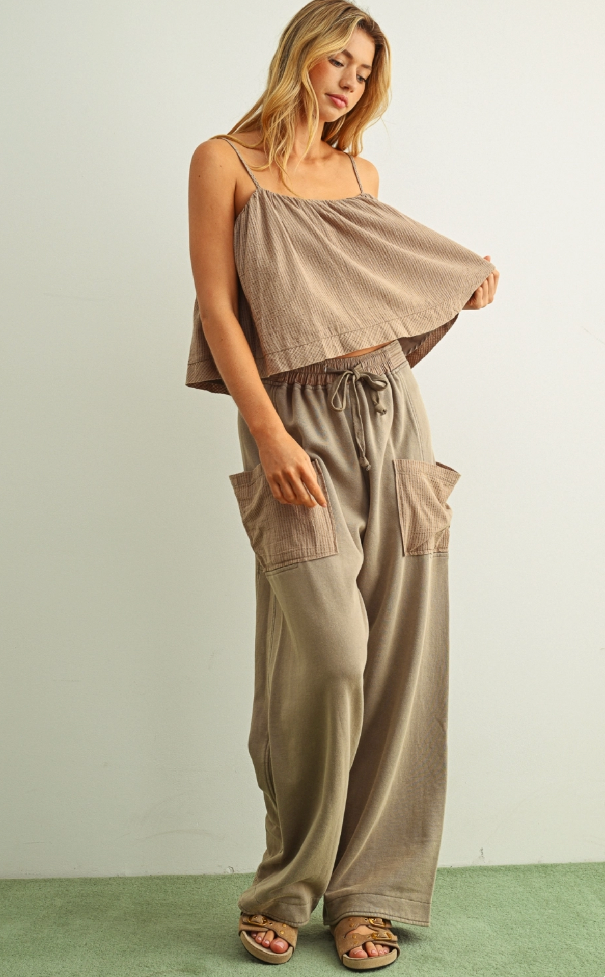 A woman modeling a two-piece lounge set consisting of a taupe off-the-shoulder crop top and matching wide-leg drawstring pants with cargo pockets, paired with brown sandals against a neutral studio backdrop.