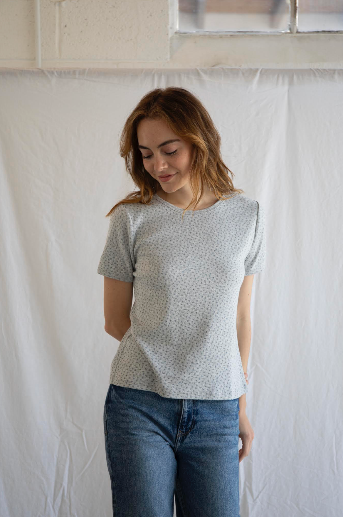 Woman wearing a light gray t-shirt with subtle pattern and blue jeans against a white curtain background