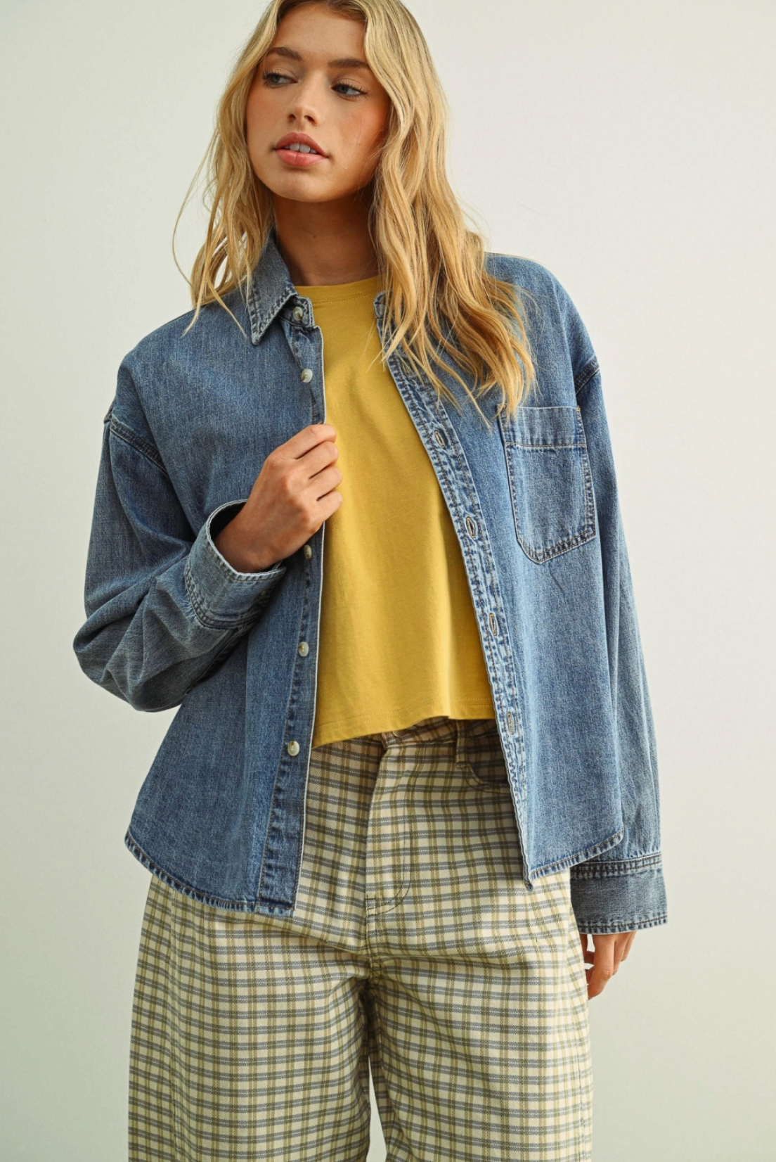 Woman wearing a denim jacket, mustard shirt, and plaid pants on a plain background