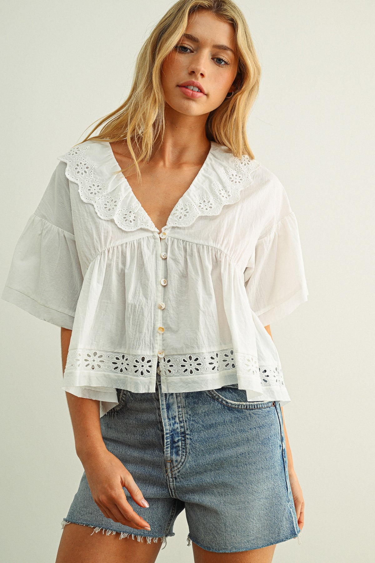 Woman wearing a white blouse with lace details and denim shorts on a plain background