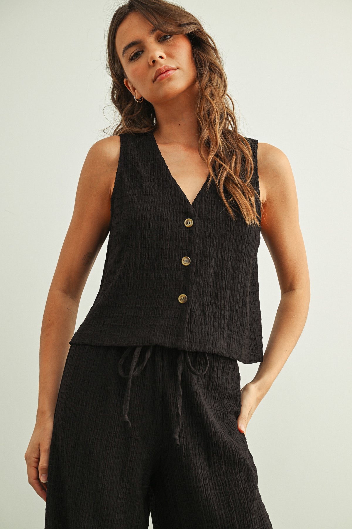 Woman wearing a black sleeveless top with buttons and black pants on a plain background