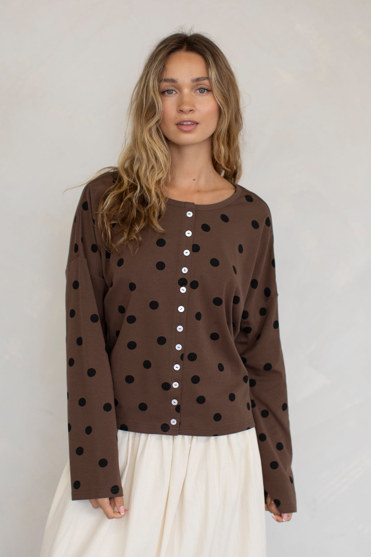 Woman wearing a brown long-sleeve top with black polka dots and white buttons on a plain background