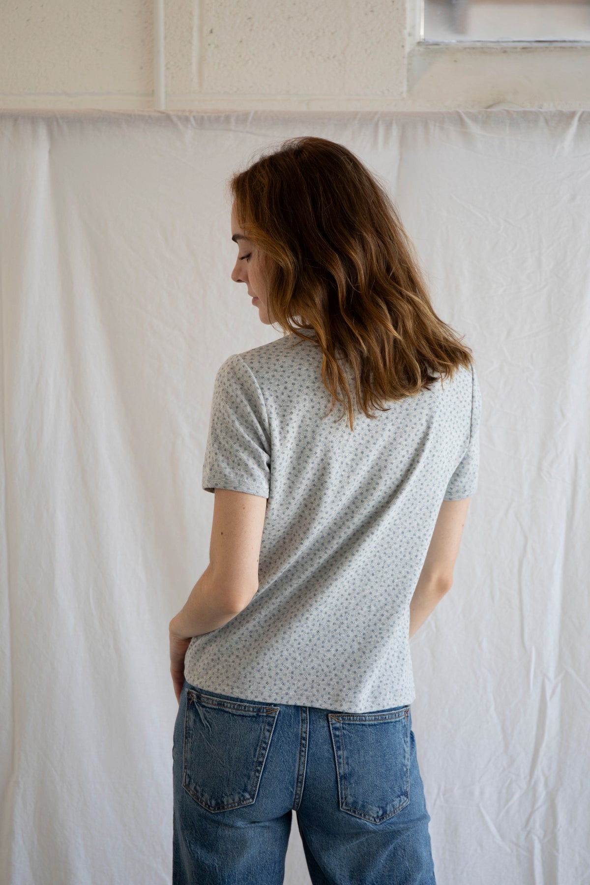 Person wearing a light gray patterned shirt and blue jeans standing against a white curtain.