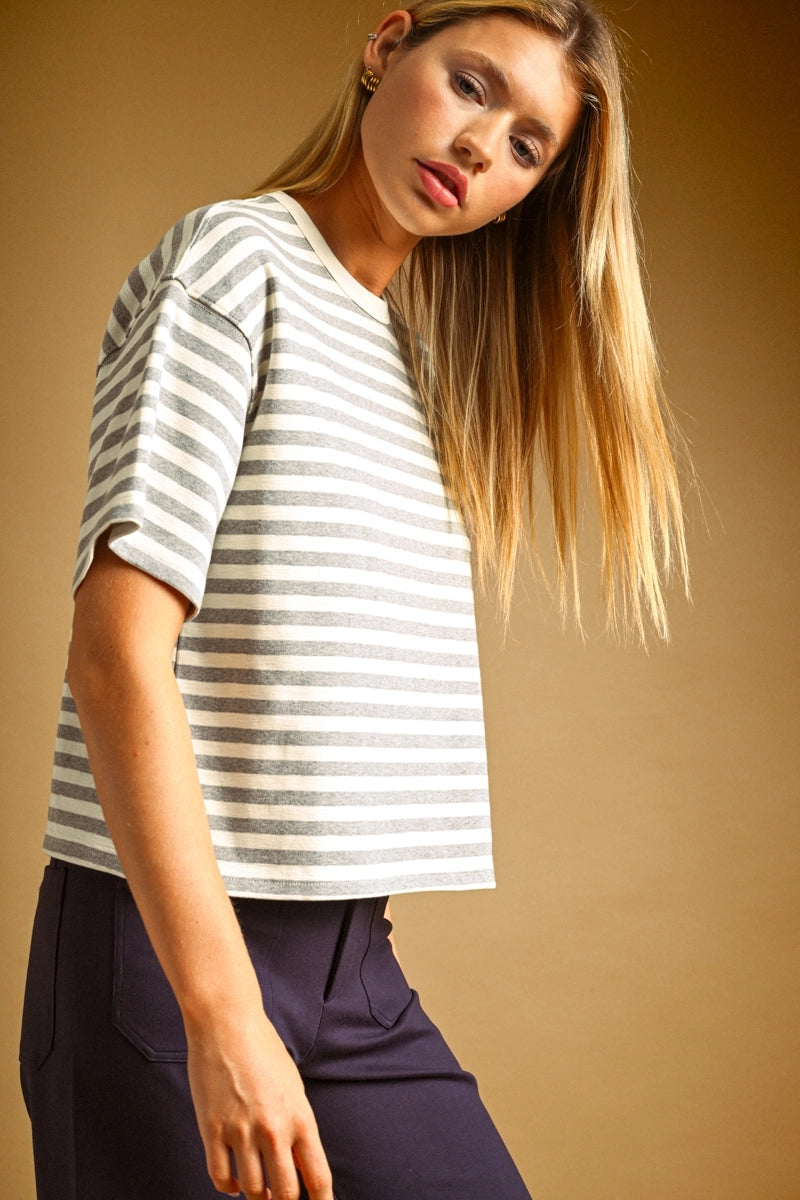 A woman wearing a short-sleeve striped top with dark navy pants, posed against a solid warm brown studio background.